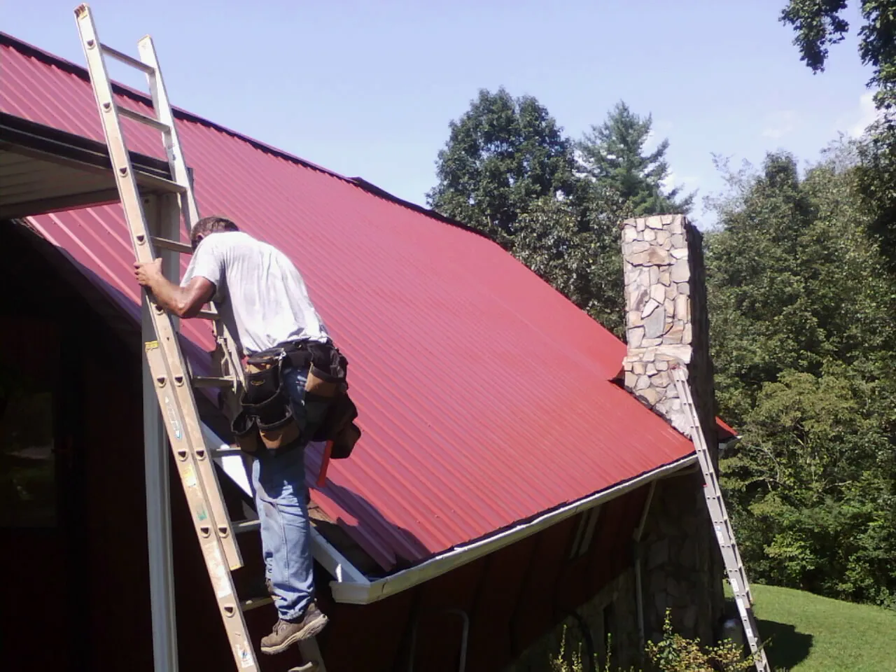 Roofer on a ladder beside a red metal roof and stone chimney — Soffit & Fascia Repair by Roofing Brothers in Index